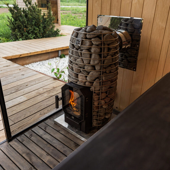 Huum Hive Flow Wood Fired Sauna Heater next to a glass wall inside of a custom sauna with a boardwalk outside on property.