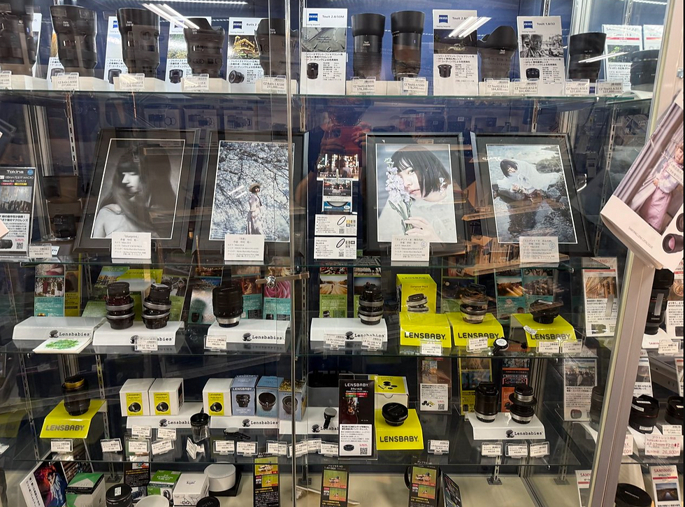 カメラ店のガラス棚に、黒いレンズやLENSBABYの箱、作例写真が並ぶ。日本語の説明は作品に使用した商品名などを記載している