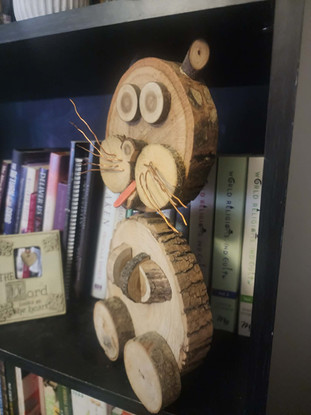 Rustic handcrafted wooden cat figurine made from natural wood slices and branches, featuring round eyes, wire whiskers, and a playful expression. Displayed on a bookshelf among books, this woodland-inspired woodworking piece highlights the beauty of natural wood grain and artisan craftsmanship.