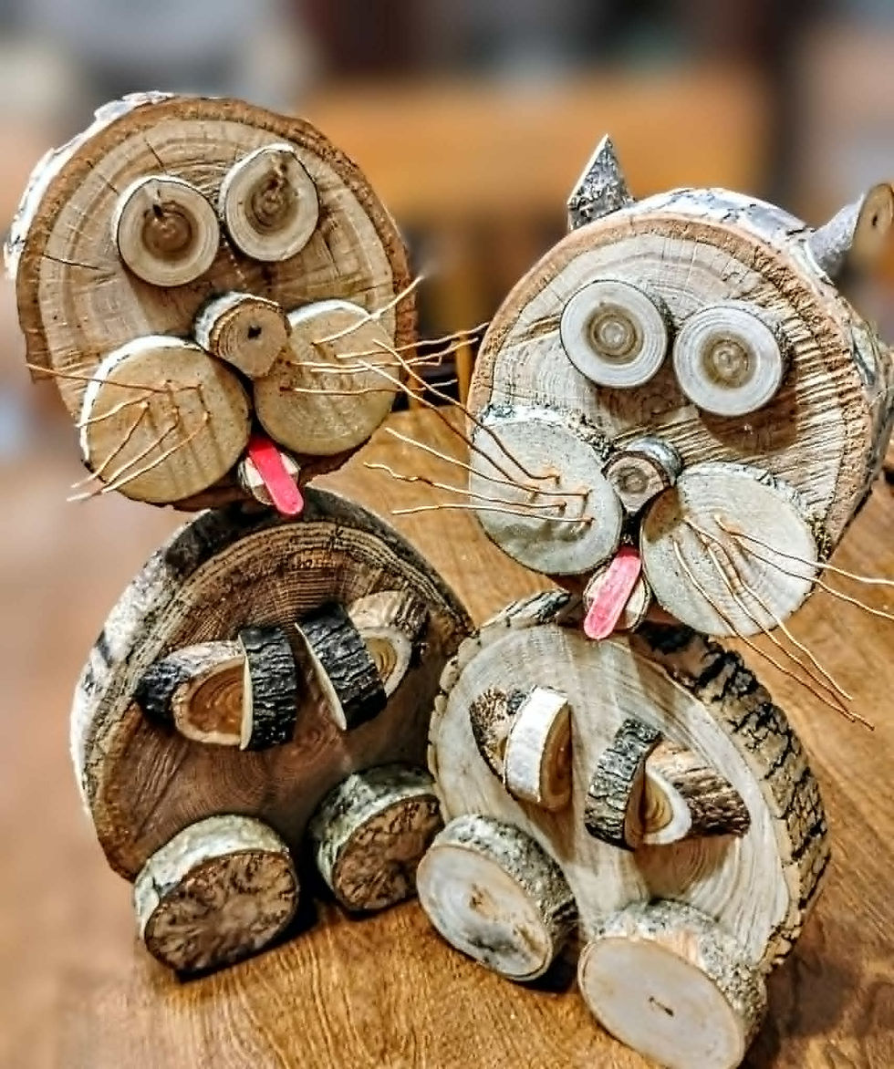 Handcrafted wooden cat figurines made from natural tree branches and wood slices, featuring rustic details such as whiskers, round eyes, and playful expressions. These unique woodland-inspired creations sit on a wooden surface, showcasing artisan woodworking and natural wood grain textures.