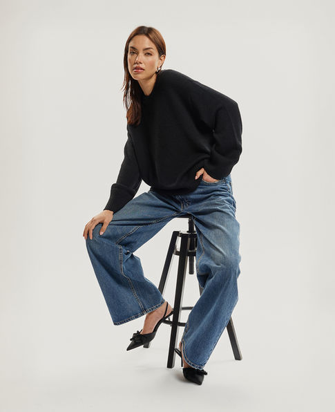 Woman wearing black sweater and blue jeans sitting on stool. E-COMMERCE/FASHION Jenia Ria.