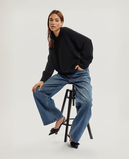 Woman wearing black sweater and blue jeans sitting on stool. E-COMMERCE/FASHION Jenia Ria.