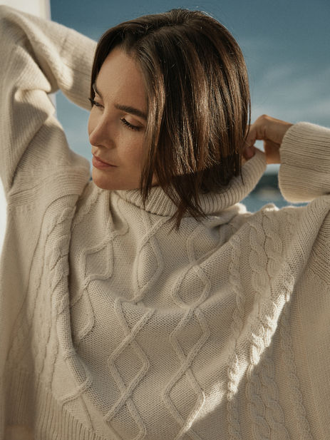 Woman in cream cable-knit sweater on a sailboat, touching her hair, campaign photoshoot for knitwear brand