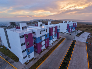 La Presidenta Claudia Sheinbaum entregando simbólicamente las llaves de una casa nueva a una familia mexicana sonriente frente a un conjunto habitacional moderno.