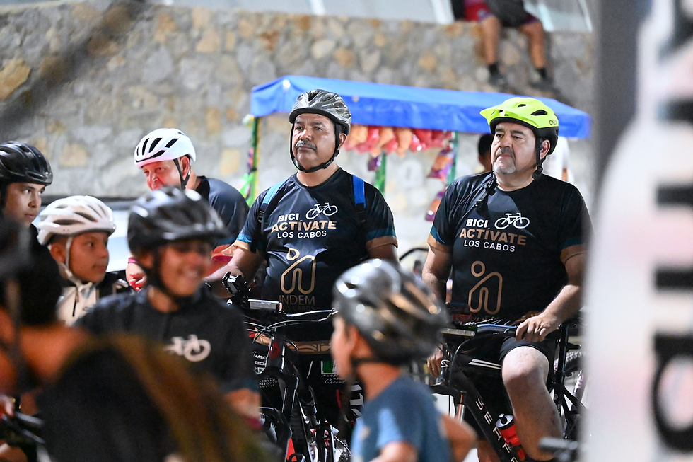 Un grupo numeroso de ciclistas de todas las edades, usando cascos y luces reflejantes, sonriendo mientras avanzan ordenadamente por la ciclovía de San José del Cabo durante el atardecer.
