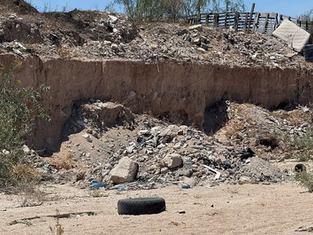 Dry creek bed in La Paz heavily polluted with plastic bags, dry overgrown vegetation, and accumulated construction debris under the sun.