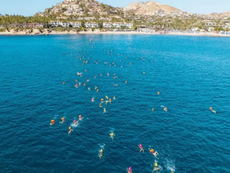 Nadadores con gorras de colores brillantes iniciando la competencia de aguas abiertas en las cristalinas aguas de Los Cabos, con el arco al fondo.