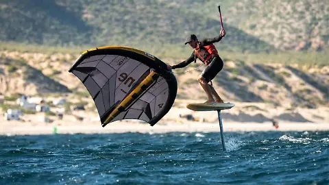 Grupo de atletas de windsurf y kitesurf navegando a alta velocidad sobre el mar azul profundo con la costa desértica de Baja California Sur de fondo.