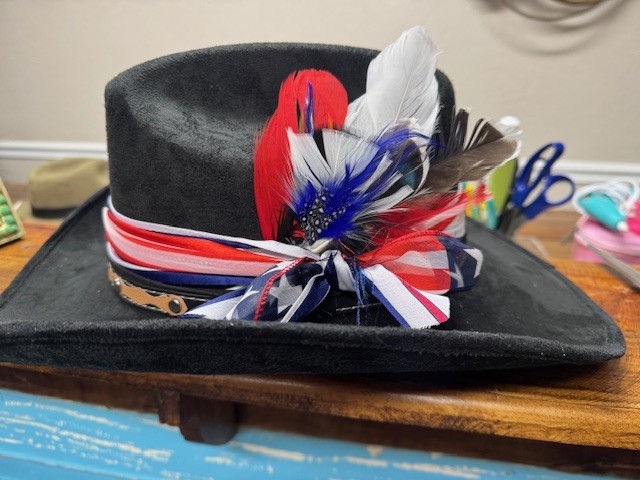 A black custom hat made of suede. Accessories include red, white, and blue ribbons, colorful feathers and a brown leather hat band.