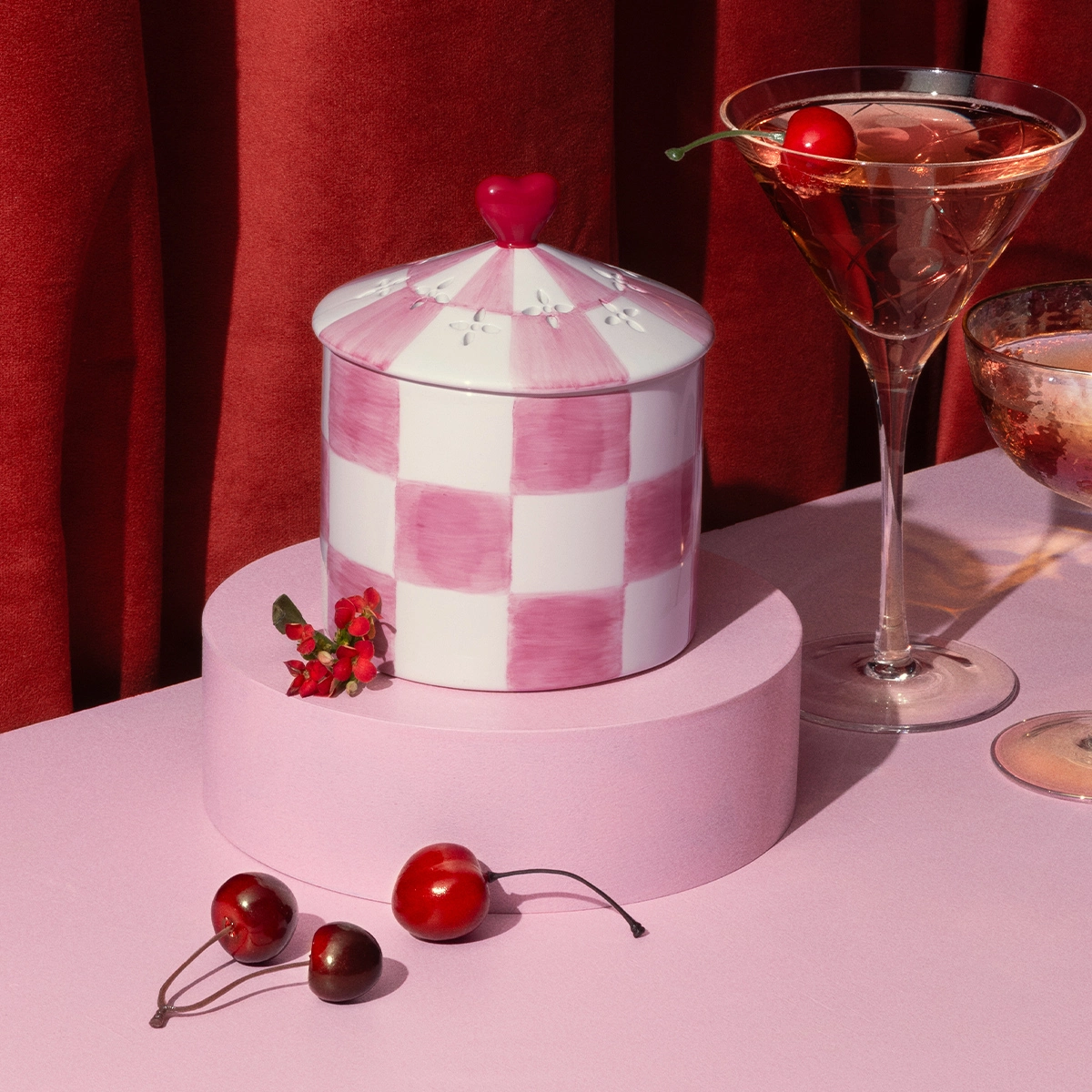 Die Scentsy Duftlampe der Valentinstag Kollektion 2026  Adonred Affair. Rosa weiß karierte Keramik. Rund mit Spitzem Deckel und einem roten Herz. Auf Rosa Podest und Rosa Untergrund mit rotem Samtvorhang im Hintergrund