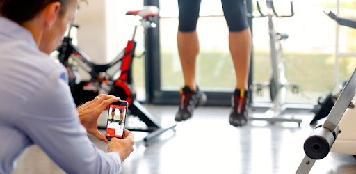 A clinician using the My Jump 2 app on a smartphone to measure a vertical jump performance in a gym setting with cycling equipment in the background.