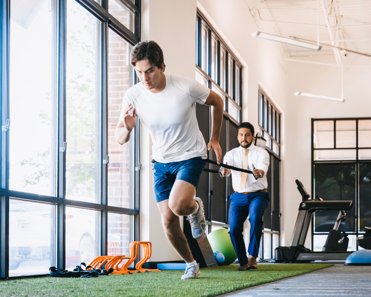 A male athlete performing sprint drills with resistance bands under the supervision of a sports medicine professional in a rehabilitation setting.
