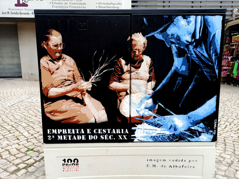 Pintura em Albufeira que homenageia a cestaria e a empreita. Um homem em azul tece um cesto (primeiro plano), e duas mulheres trabalham na empreita ao fundo, recriando o trabalho manual e comunitário da região.