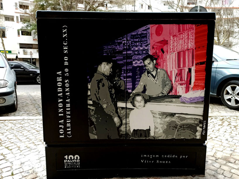 Pintura "Homenagem ao Comércio Histórico" terminada num armário de electricidade em Albufeira. Figuras (vendedor, cliente, criança) pintadas em tons de cinza (nostalgia), contrastando com os tecidos vibrantes (violeta, rosa) da antiga Loja Inovadora. Técnica Stencil e Spray.