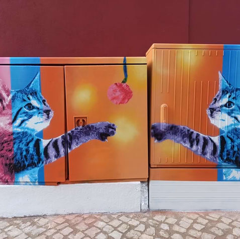Pintura em Stencil e Spray num armário de electricidade em S. B. Messines. A obra retrata dois gatinhos e um novelo em tons vibrantes de azul, violeta e rosa, sobre um fundo laranja forte e contrastante, num estilo pop e lúdico.