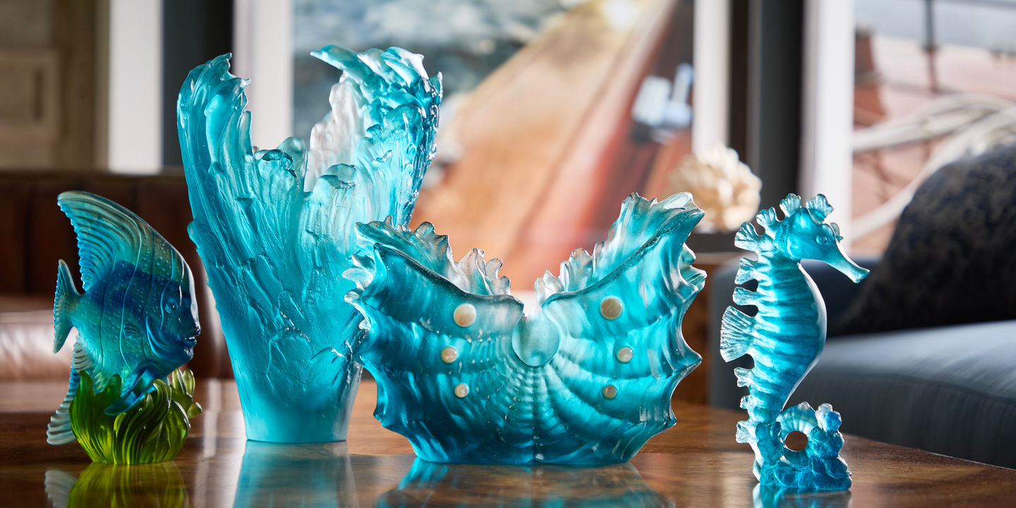 The image shows a collection of beautiful blue glass sculptures. There's a large wave-shaped vase, a smaller seashell-shaped bowl, a fish, and a seahorse. All the pieces are made of clear blue glass and have a translucent quality.