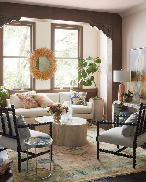The image shows a cozy living room with a warm and inviting atmosphere. There's a large, comfy sofa with colorful throw pillows, two armchairs, and a coffee table made of natural stone. A large sunburst mirror hangs on the wall, and a colorful rug covers the floor. The room has large windows that let in plenty of natural light.