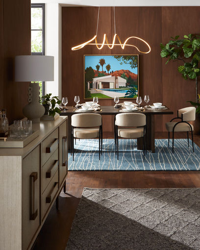 The image shows a stylish and modern dining room with a warm and inviting atmosphere. The room features a dark wood wall, a large rectangular dining table with beige upholstered chairs, and a unique curved light fixture hanging above. A colorful painting of a house with a pool hangs on the wall, adding a touch of vibrancy. The room also includes a wooden sideboard and a patterned rug, completing the stylish and contemporary look.