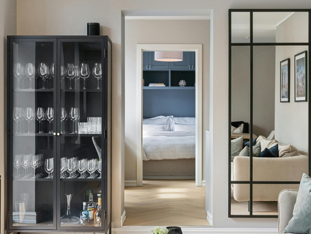 Glass cabinet with wine glasses, a cozy bed in a blue-walled room visible through a doorway, a beige sofa with cushions on the right. Organized downsized condo.