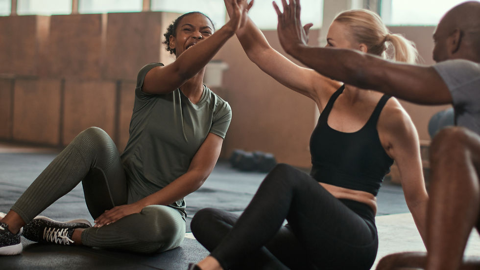 Three people in workout gear sit on a gym floor, high-fiving and smiling. Large windows in the background. Positive and energetic mood.
