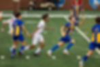 Soccer players in blue-yellow uniforms chase the ball on a green field. Others in white run nearby. Background includes a cart and brick wall.