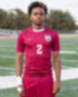 A soccer player stands on a field wearing a maroon jersey with number 2.Trees in the background.
