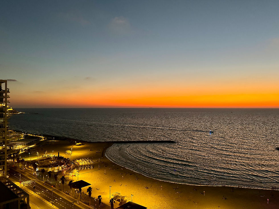Tel Aviv beach at sunset, with the Mediterranean Sea stretching toward the horizon, city lights illuminating the shoreline, and calm waves reflecting the fading light.