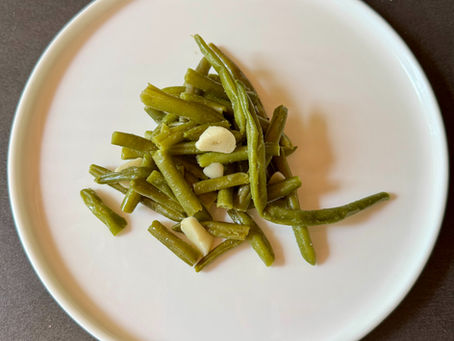 Assiette blanche vue du dessus contenant une petite portion de haricots verts cuits, avec quelques éclats d’ail, posée sur un fond sombre.