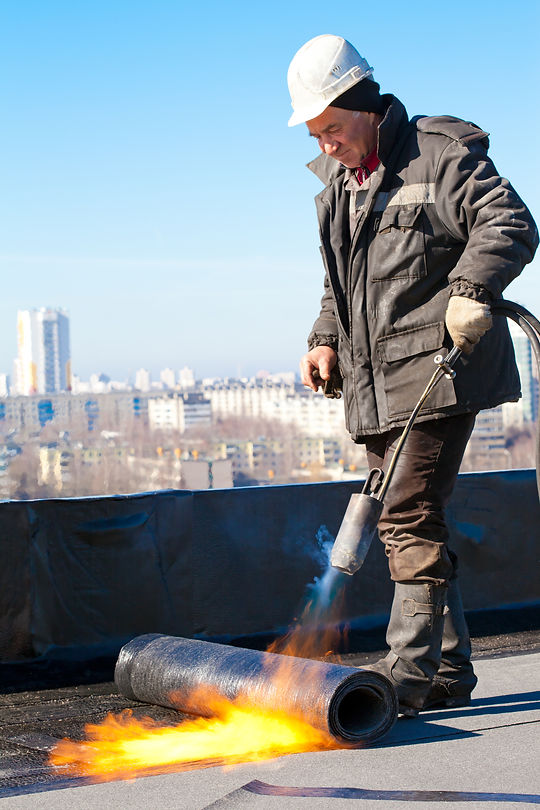 Roofer man worker in helmet installing a roll of roofing felt with gas blowpipe torch during constru