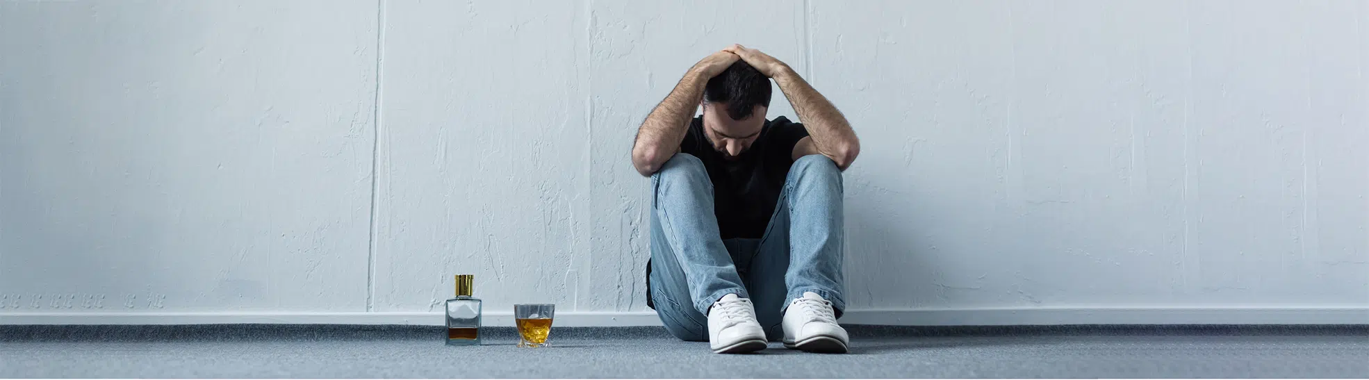Panoramic shot of suffering man sitting on floor by white wall near bottle and glass of whiskey copy