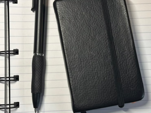 A black notebook bound with an elastic sitting on lined paper beside a black ballpoint pen