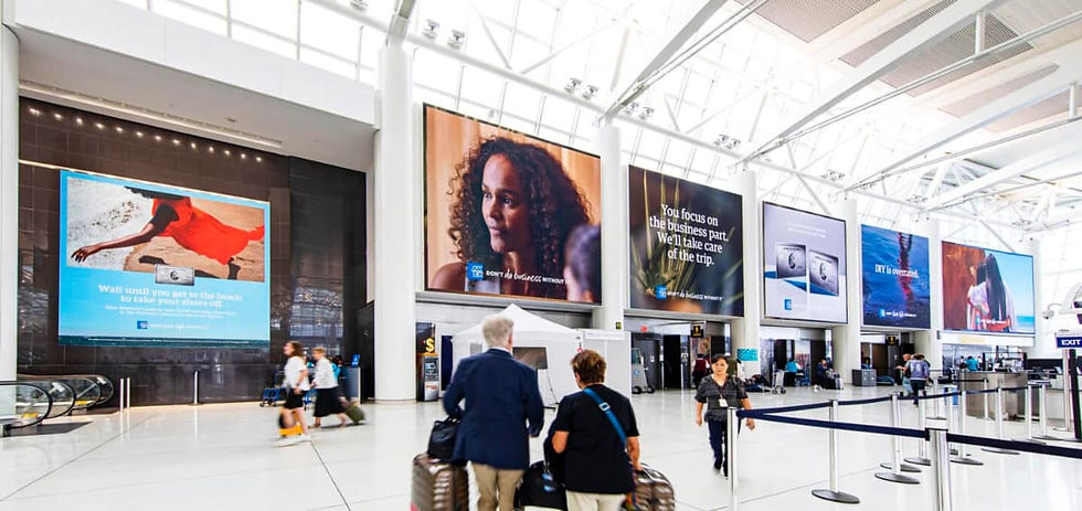 People walk and sit in a shopping area with tech ads on digital screens. The ceiling has modern lights; shops line the background.