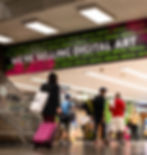 People walk in a subway station with luggage. A large banner reads "WE'RE SELLING DIGITAL ART." Bright pink and green accents.