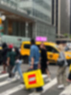 People crossing a street in front of Bank of America Tower. Background includes yellow taxis with digital top advert dispay