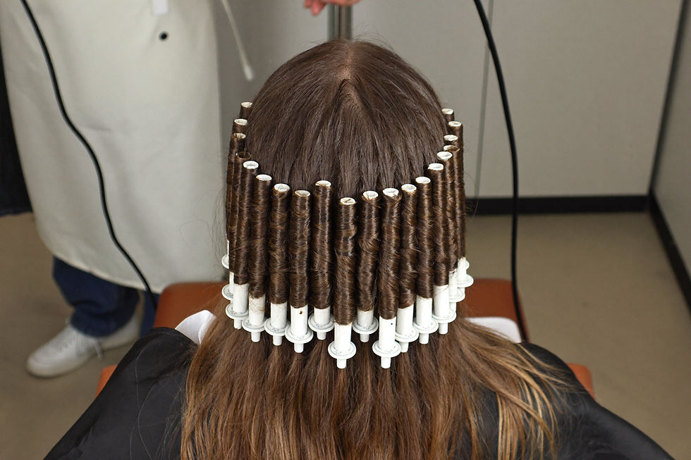 Back of a seated client head with long hair wound onto rows of perm rods hanging downward during a spiral perm service