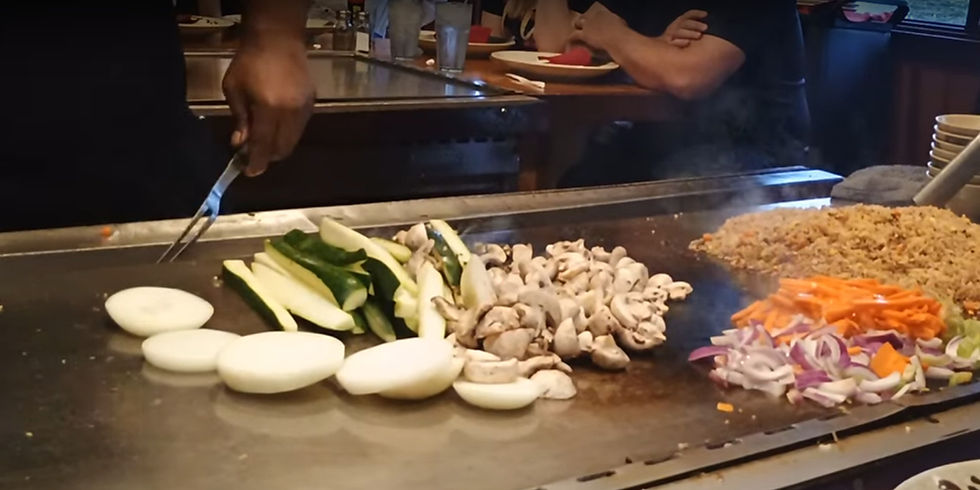Chef cooking onions, zucchini, mushrooms, and fried rice on a hibachi grill. Diners watch in background, colorful vegetables in foreground.