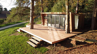 Wooden cabin with decking and steps infront in a garden, surrounded by trees