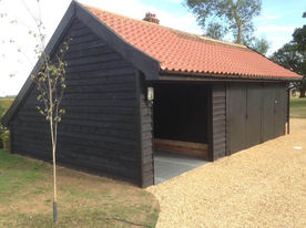 A black wooden timber garage with an orange tiled roof, featuring one open parking bay and closed double doors on a gravel driveway.