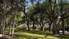 **Alt text:** Large sloping front lawn shaded by mature oak trees, with a light stone home visible at the bottom of the hill in Central Texas.