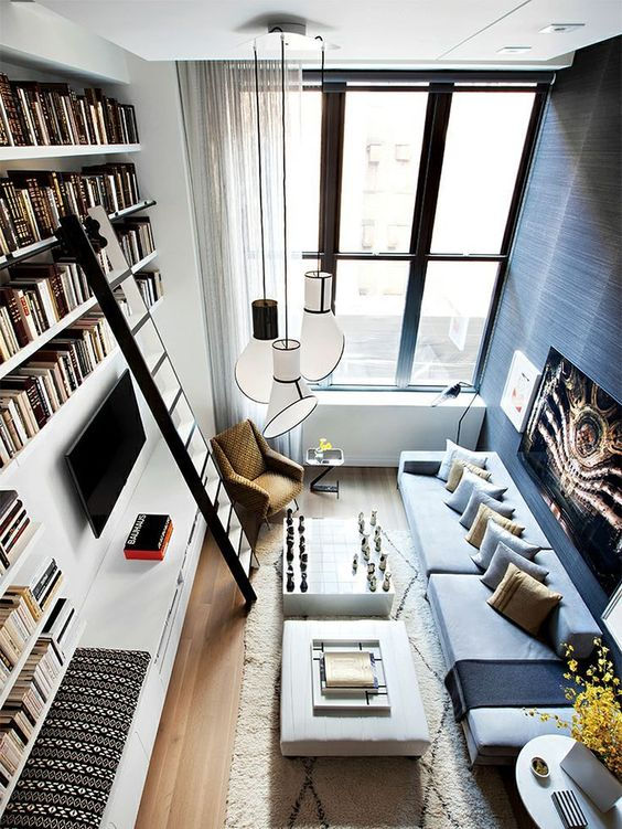 Bibliothèque toute hauteur, peinte en blanc comme les murs, et parquet à chevrons au sol. Fauteuil en bois et assise et dossier gris.