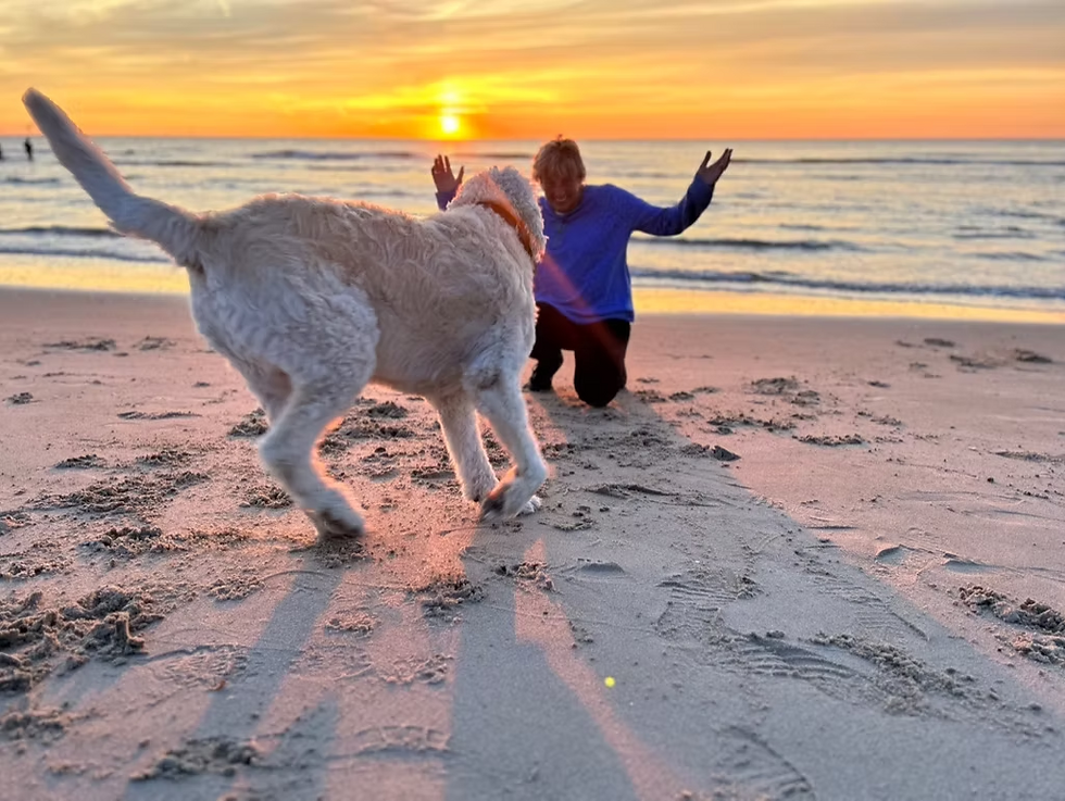 Yvonne van der Meer speelt met haar Labradoodle Buddy op het strand bij zonsondergang – symbool voor vertrouwen, plezier en een sterke band tussen hond en eigenaar