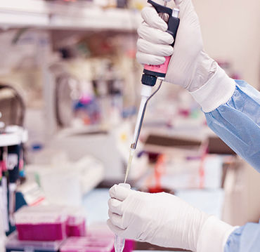 Person in blue lab coat using pipette in a laboratory. Gloves are worn, pink liquid in tray, blurred shelves in background. Clinical setting.