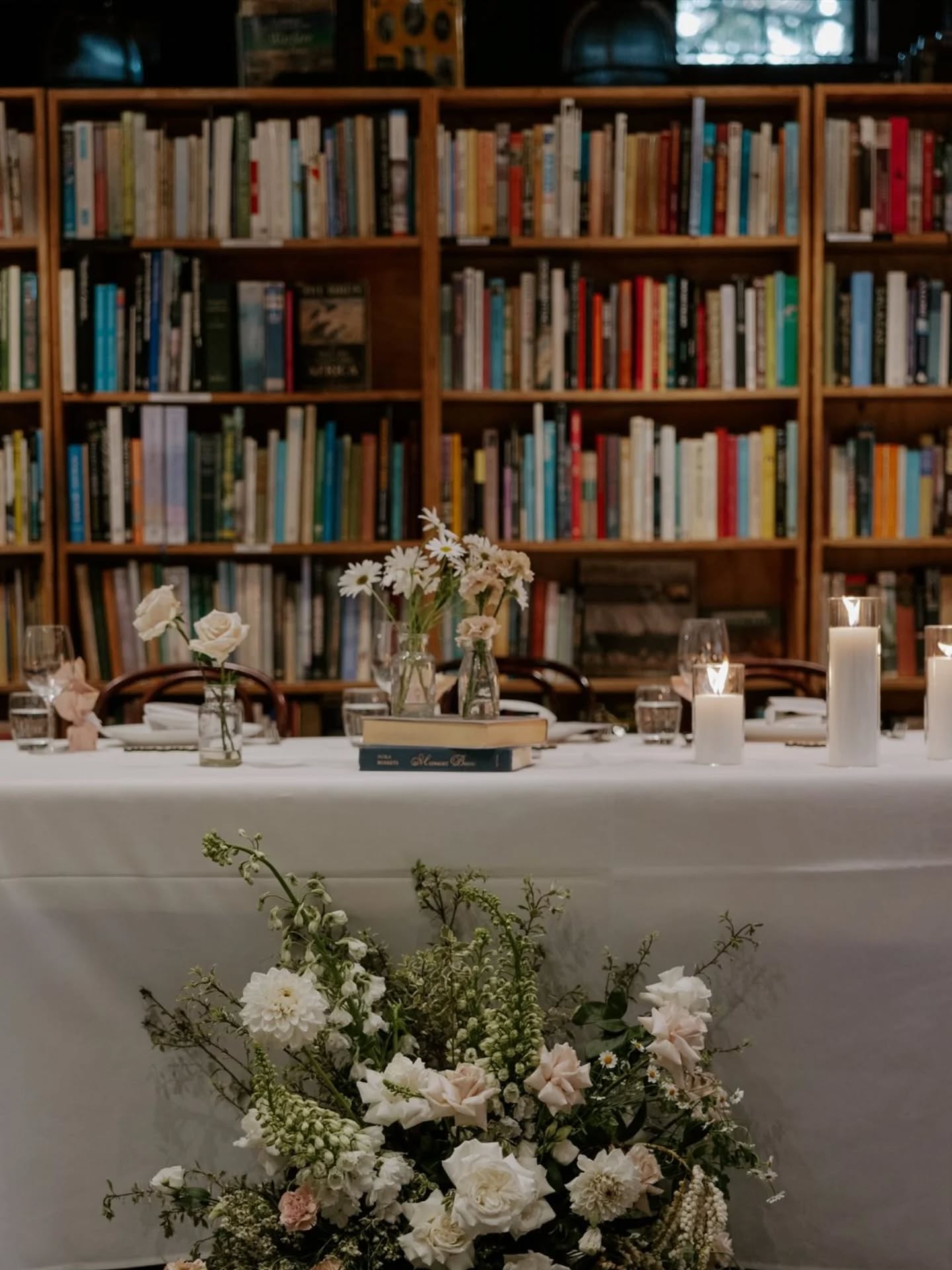 bridal table with bookshelf behind, on table books scattered with bud vases of flowers