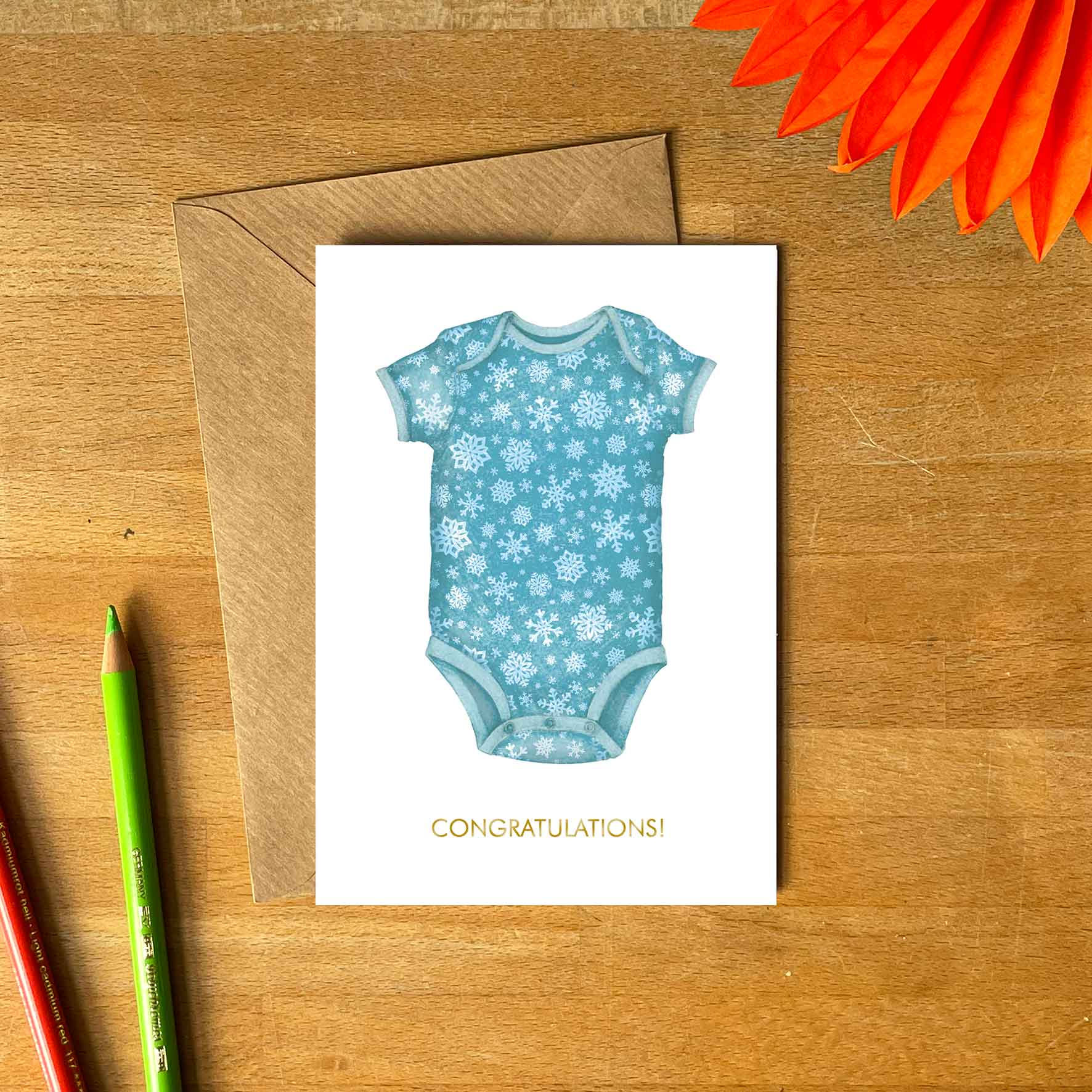 white greetings card with blue baby romper with snowflakes pattern and congratulations text underneath, kraft envelope
