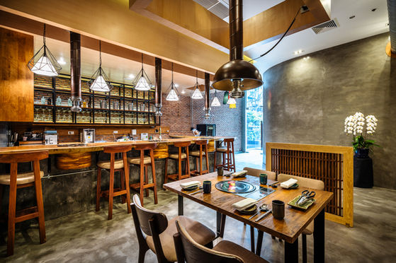 Modern industrial dining space featuring a live-edge wooden bar, brick walls, geometric pendant lights, and a table set for grilling with warm wood furniture and concrete flooring.
