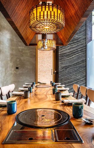 Elegant private dining room with a long live-edge wooden table, integrated grill stations, cushioned chairs, warm decorative lighting, and a mix of concrete and stone wall finishes.
