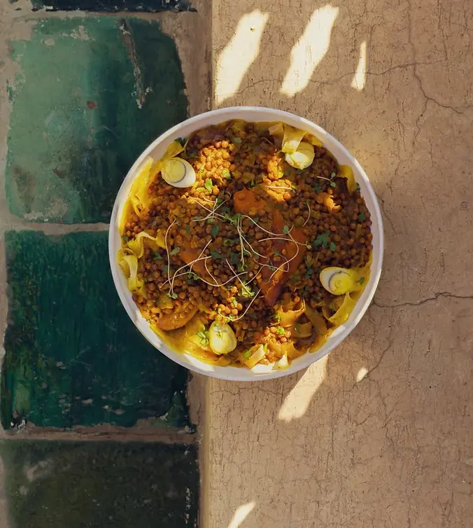 Overhead view of a bowl of lentils and pasta with egg slices and herbs on a sunlit cracked wall, warm rustic feel