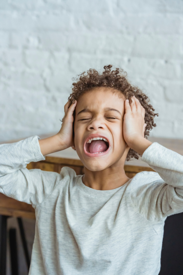 A boy in a white shirt having a meltdown. He is holding his head, his eyes are closed, and his mouth wide open screaming.