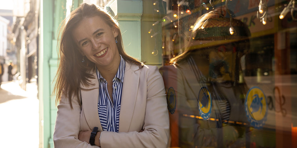 Rachel smiling at the camera, dressed in a blue striped shirt and a business jacket leaning against a window