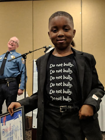 Young man smiling who won an award wearing a t-shirt that says "Do not bully", with a police chief smiling in the background.